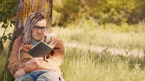 Ein Mann in Freizeitkleidung und Brille liest Literatur unter einem Baum