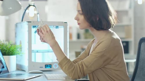 Young Woman Engineer Expects 3D Printed Part that She Just Made with Her 3D Printer.