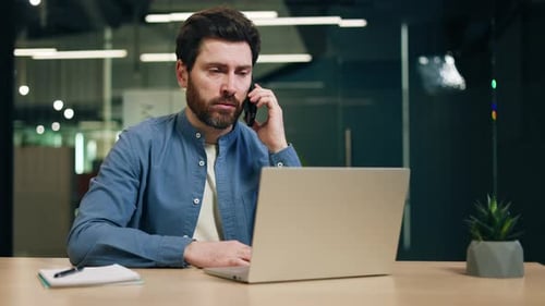 Busy Man Talking on Smartphone While Sitting in Office at Desk with Computer