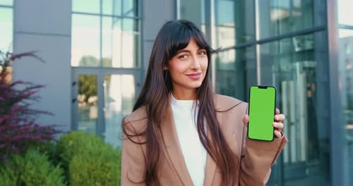 Professional young businesswoman in formal attire holding a smartphone with a green screen in a mode