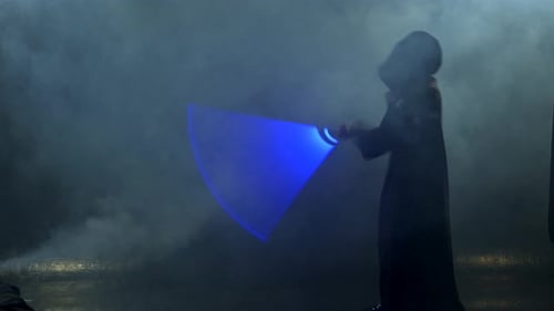 Young Man in Jedi Cosplay Costume with Lightsaber Battle on Black Background in Smoke and Rain Slow