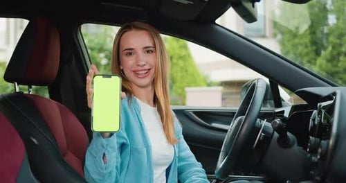 Happy smiling woman driver sitting behind the wheel of premium modern car showing smartphone