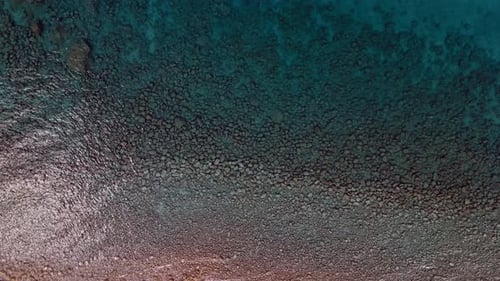 An aerial drone shot of of a crystal clear ocean shoreline with turquoise water and small pebbles.