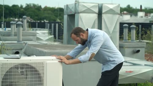 Young man with laptop inspecting air conditioning unit on city rooftop
