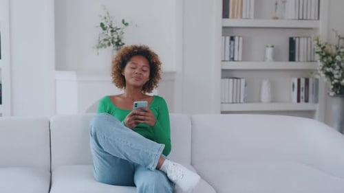 Young Optimistic Pretty African American Woman with Phone in Hands Sits on Sofa