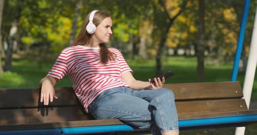 Woman in Wireless Headphones Swinging on Swing Listening to Music in City Park