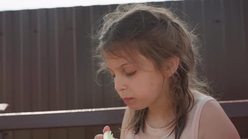 Une fille caucasienne découvre la saveur du melon avec un regard concentré sur le banc du porche, des bouchées délicates et