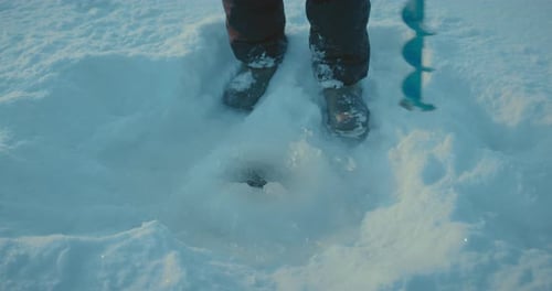 Der Mensch bohrt ein Eisloch in einem gefrorenen Fluss Nahaufnahme von Fisher Shoes und Ice Drill Prores