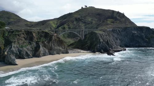 Drone flies over the water showing a beautiful beach beneath one of the iconic bridges along highway