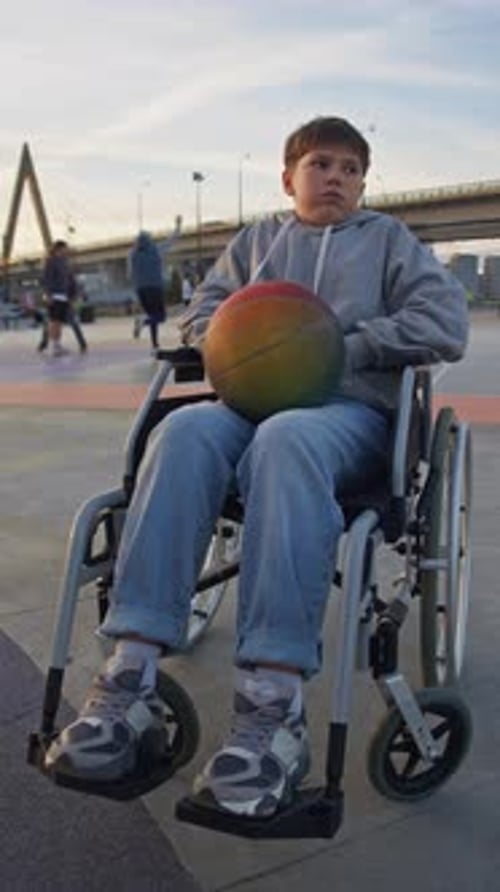 A Lonely and Upset Boy in a Wheelchair Holding a Basketball Sits Against the Backdrop of a Street