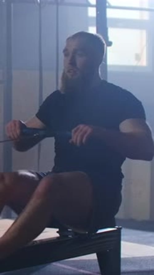 Bearded Athlete Powering Through an Intense Indoor Rowing Machine Workout in a Dimly Lit Gym