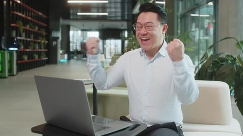 Chinese Young Businessman in Shirt Working on Laptop Shocked By Sudden Victory in Modern Hotel Lobby