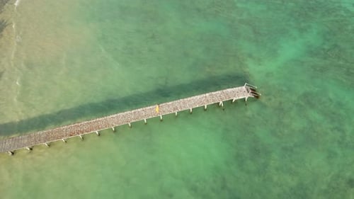 A pessoa caminha em um píer de madeira envelhecido que se estende até águas esmeraldas rasas com fundo marinho visível