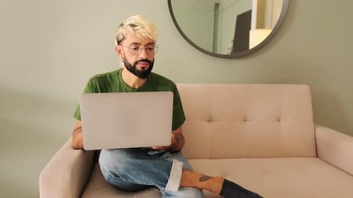 Young blond man with a beard works on a laptop while sitting in a cozy living room