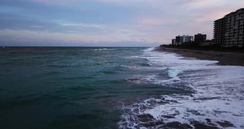 Drone footage. Aerial of coastline along shallow surf, with view of coastal city buildings.