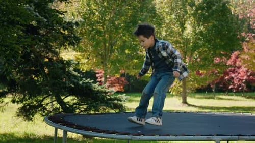 Happy Boy Jumping on a Trampoline Outdoors Surrounded By Green Trees and Autumn Colors Enjoying an