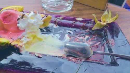 A closeup view of the tray of eaten ice cream and fruits with a spoon on the table.
