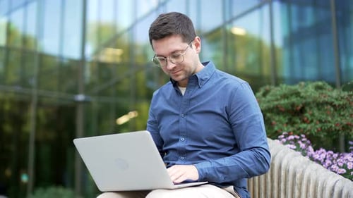 Confident businessman works on a laptop computer sitting on a bench near business office building.