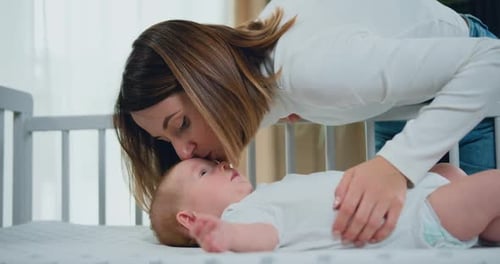 Молодая мать обнимает и целует свою очаровательную многомесячную малышку, отдыхающую в кроватке и улыбающуюся.