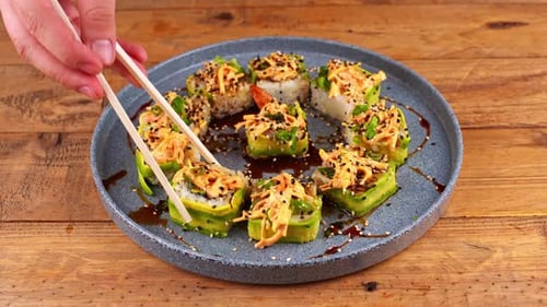 Close-up of hands with chopsticks, awkwardly picking up an avocado sushi roll from a blue plate.