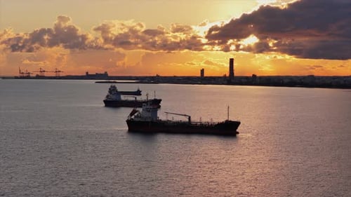 Aerial drone view of multiple oil tankers anchored near an industrial port at sunset with cranes and