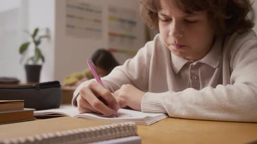 Boy Sitting at Desk in School and Writing in Notebook