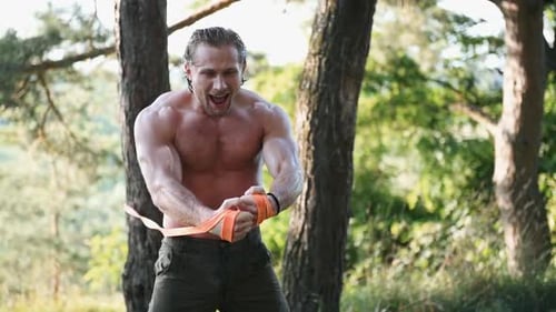 Strong shirtless man with muscular body type doing workout in the forest by using fitness resistance