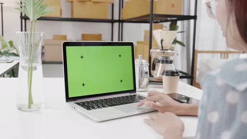 Over the Shoulder : Young Woman Sitting at Her Desk Using Laptop with Mock-up Green Screen.