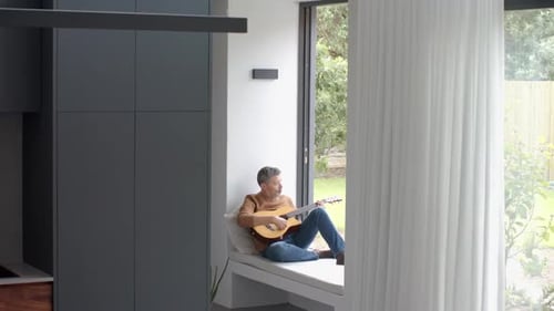 Man Playing Guitar on Window Bench, Relaxing at Home