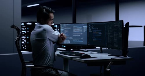 Side View Of Asian Male Programmer Stretching While Writing Code By Desktops In The Server Room