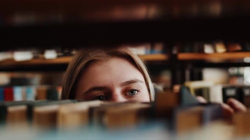Junge kaukasische Studentin steht zwischen Bücherregalen und wählt sorgfältig ein in ihr verwendetes Buch aus