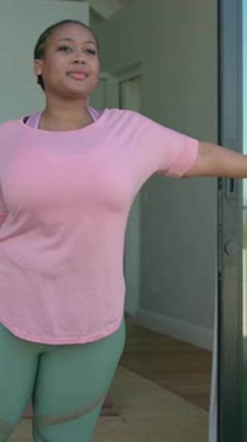 Vídeo vertical: batente de porta, mulher em camiseta rosa, leggings de sálvia estendendo os braços e levantando os joelhos