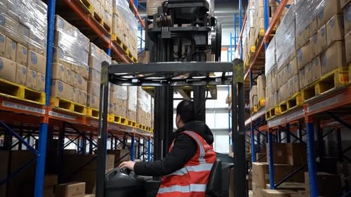 Warehouse worker driver in uniform forklift riding between racks storehouse with pallet of cardboard