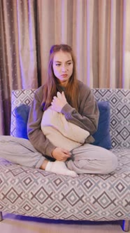A Sad Young Woman Sits Alone on a Couch Hugging a Pillow Tightly Lost in Her Thoughts A Melancholic