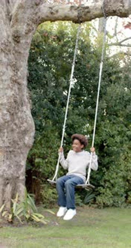 Vertical video of happy biracial boy swinging on tree swing in garden, in slow motion