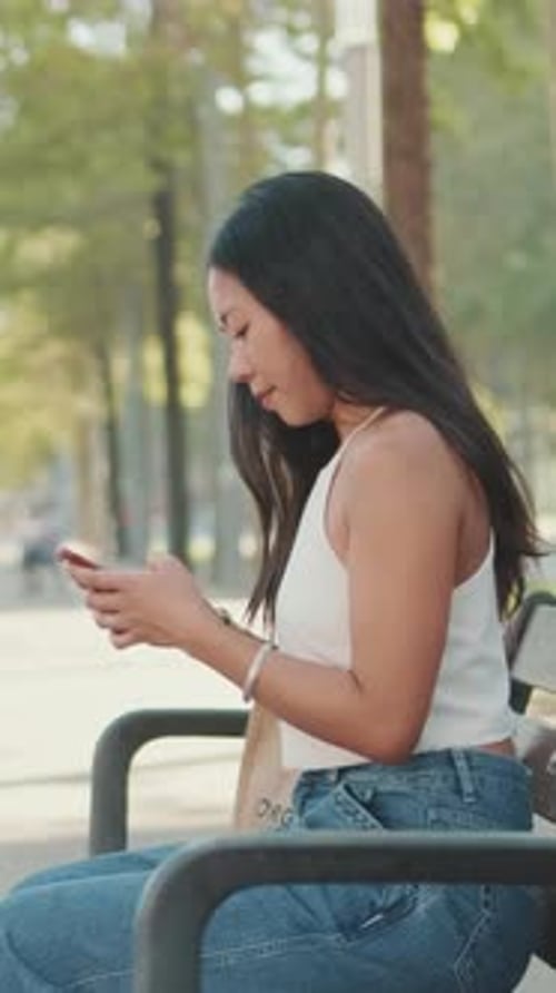 Young woman sitting on park bench typing message on mobile phone