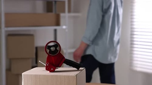 Man using dispenser with adhesive tape to seal cardboard box indoors, closeup