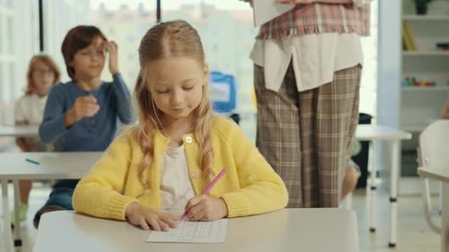 Beautiful Longhaired Blonde Pupil Sits at a Desk at School and Fills Out Tests and the Teacher