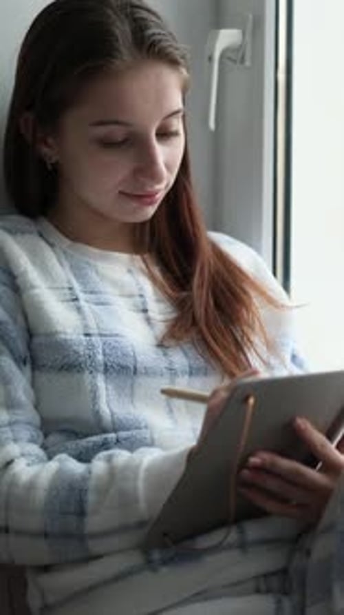 Teenage Girl Sitting On Windowsill Reading Notes In Notebook