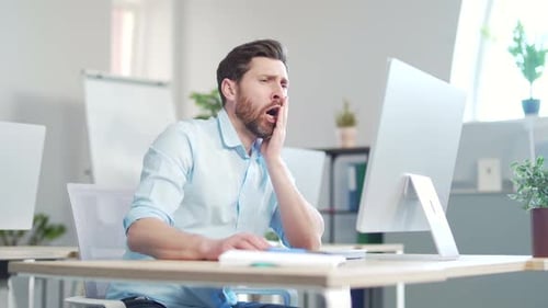 Bored sleepy office worker sitting at work workplace at computer desk leaning on hand yawning. Tired