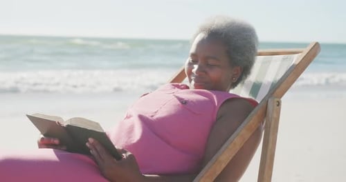 Happy senior african american woman sitting on deck chair and reading book at beach, in slow motion