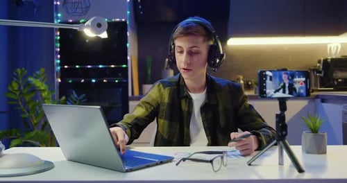 Chico guapo bloguero de redes sociales con auriculares mira la pantalla del portátil y pronuncia un discurso