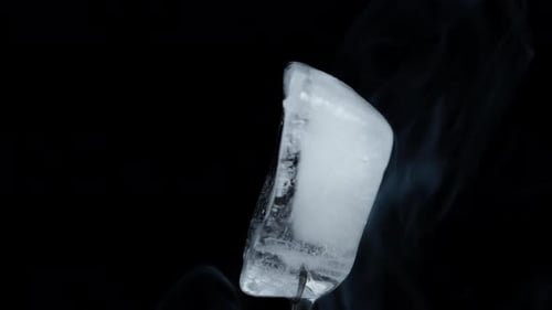 An ice cube rotates on a black background, emitting smoke, in a close-up shot. Isolate