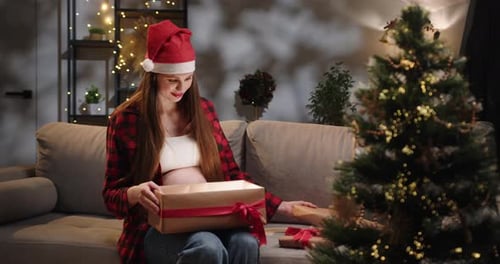 Pregnant Woman Opening Christmas Gift by Tree Indoors