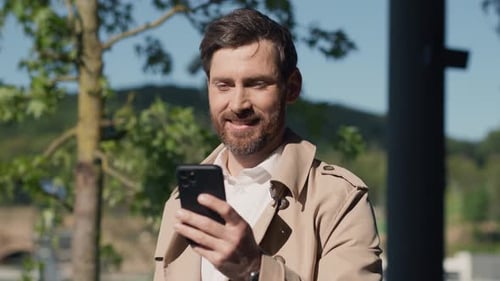 At Sunlight Happy Smiling Stylish Handsome Man with Beard in Coat Uses Smartphone Stand in Part