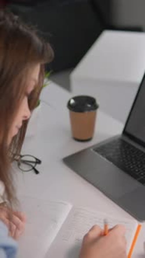The Diligent Student at the University Desk is Fully Focused on Taking Detailed Notes Using a Laptop