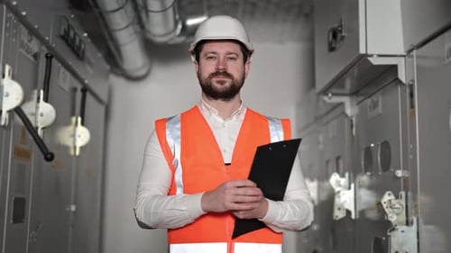 Electrical Engineer in Orange Vest and White Helmet with Clipboard in Control Room