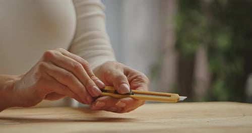 Hands Holding a Yellow Utility Knife on Wooden Surface