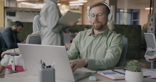 Joyful Young Employee Listening to Music with Headphones and Using Laptop in Coworking Center