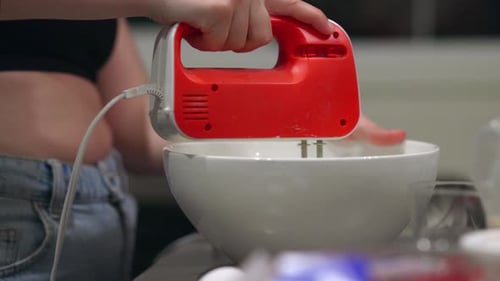 woman using a red hand mixer to mix cheesecake ingredients in a white bowl in a kitchen.